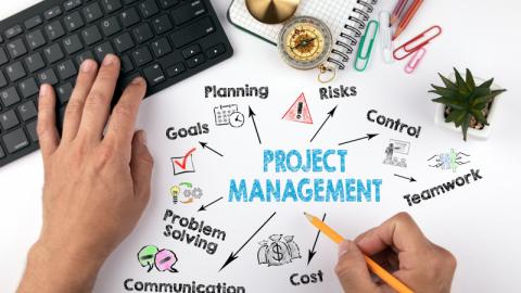 Hands on a tabletop one on keyboard one holding a pencil. The words Project Management and other related words appear on the to be being drawn.